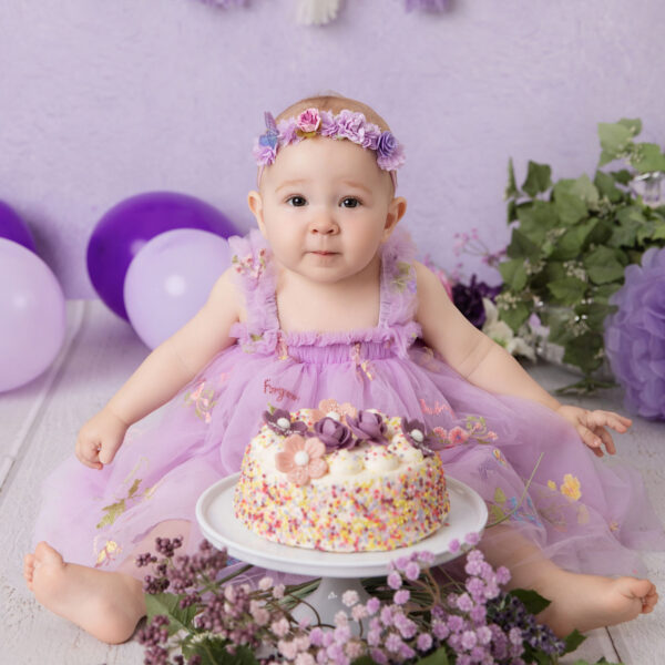 Baby im lila Kleid sitzt vor lila Hintergrund mit Mini-Torte beim Cakesmash und schaut in die Kamera, fotografiert im Studio von Belly Deluxe in Karlsruhe.