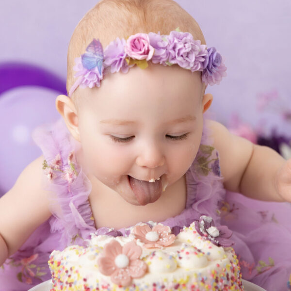 Nahaufnahme eines Babys im lila Outfit mit Blumenkranz, das beim Cakesmash die Zunge rausstreckt vor lila Hintergrund, fotografiert im Studio von Belly Deluxe in Karlsruhe.