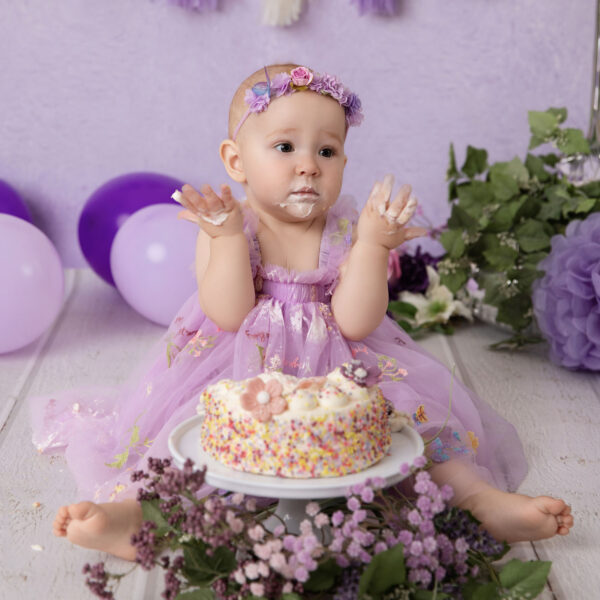 Baby im lila Kleid zeigt beim Cakesmash cremige Hände vor lila Hintergrund mit Mini-Torte, fotografiert im Studio von Belly Deluxe in Karlsruhe.
