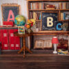 Einschulungs-Fotoshooting-Hintergrund im Klassenzimmer-Look mit Bücherregal, Tafeln mit A/B/C, rotem Spind, Globus und bunter Schultüten-Girlande.