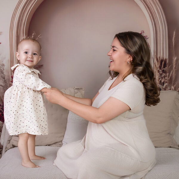 Einjähriges Mädchen steht an den Händen ihrer lachenden Mama auf dem Liegesofa im Boho-Fotoset