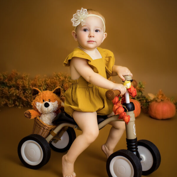 Baby im gelben Outfit sitzt auf einem Rutschfahrzeug im herbstlichen Studio-Set, mit Kürbissen und herbstlicher Deko vor warmem Hintergrund.