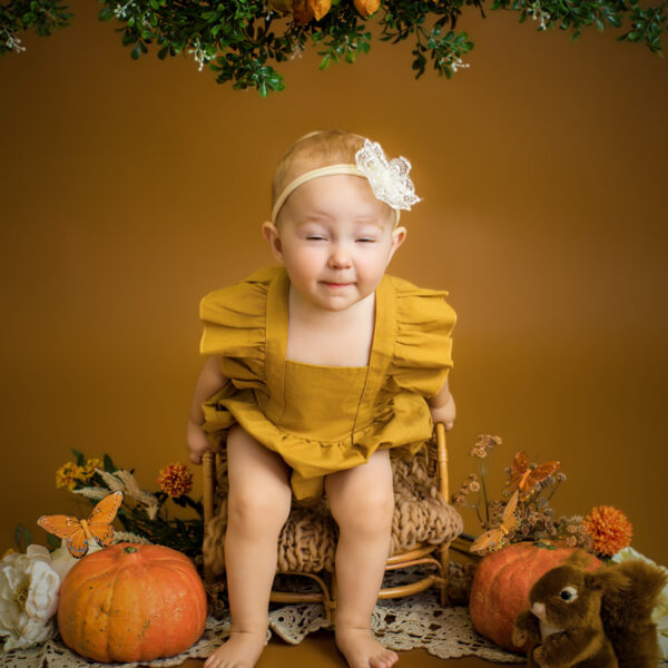 Kleinkind im gelben Outfit sitzt im herbstlichen Studio-Set auf einem kleinen Rattan-Sessel und macht eine lustige Grimasse, umgeben von Kürbissen und herbstlicher Deko vor warmem Hintergrund.