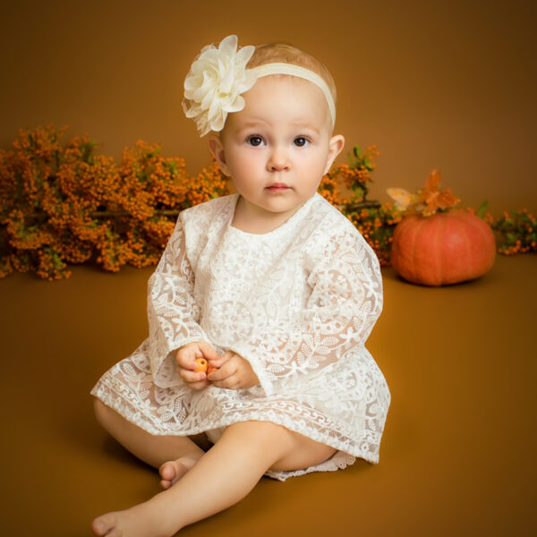 Kind im weißen Spitzenkleid sitzt im herbstlichen Studio-Set vor warmem Hintergrund, mit Kürbis und herbstlicher Deko im Hintergrund.