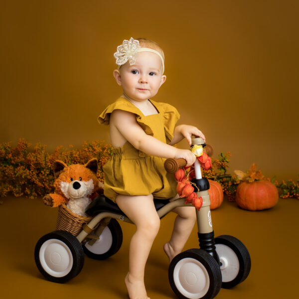 Kleinkind im gelben Outfit sitzt auf einem kleinen Rutschfahrzeug im herbstlichen Studio-Set, mit Kürbissen und herbstlicher Deko vor warmem Hintergrund.